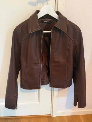 Brun skinnjacka från Gina Tricot - Snygg brun skinnjacka från Gina Tricot med klassisk krage och dragkedja framtill. Jackan har raka ärmar med slits vid ärmslut och markerade sömmar för en cool look. Perfekt till jeans eller kjol för en trendig outfit.