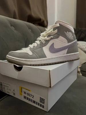 Nike Air Jordan 1 Mid grå/vit/blå - Snygga Nike Air Jordan 1 Mid sneakers i grått och vitt med klassisk siluett och ikonisk Swoosh på sidan. Skorna har högt skaft, vita skosnören och perforerad tåbox för extra ventilation. Perfekta för dig som gillar streetwear och sportig stil.