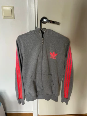 Grå Adidas hoodie med rosa ränder - Snygg grå hoodie från Adidas med dragkedja, huva och kängurufickor. Ikoniska rosa ränder längs ärmarna och Adidas logga på bröstet. Perfekt för en sportig och avslappnad stil.