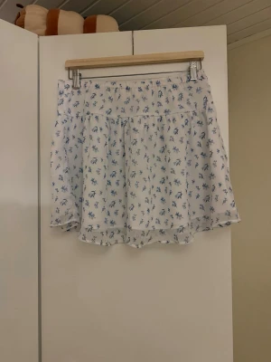 Vit kortkjol med blått blommönster - Söt vit kortkjol med små blå blommor och lätt volang nedtill. Kjolen har en luftig och flowy passform, perfekt för varma dagar. Tillverkad i ett tunt och mjukt material som känns bekvämt mot huden.