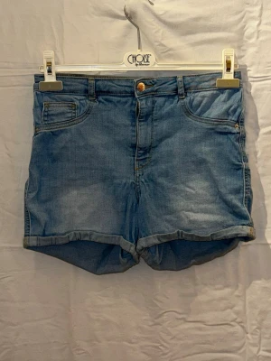 Ljusblå denimshorts med upprullad kant - Säljer ett par ljusblå denimshorts med upprullad kant och klassisk femficksdesign. Shortsen har knapp och dragkedja framtill samt bälteshällor i midjan. Perfekta för varma dagar och enkel att matcha med olika stilar.