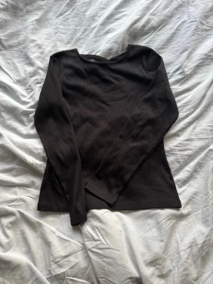 Svart ribbad långärmad topp - Snygg svart ribbad topp med långa ärmar och cool detalj med korsade band i halsringningen. Perfekt att styla med jeans eller kjol för en trendig look. Materialet känns mjukt och stretchigt.