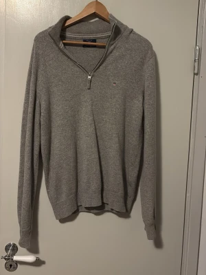 Grå stickad halvzip-tröja från Gant - Snygg grå stickad tröja från Gant i superfin lammull. Tröjan har halv dragkedja, ribbade muddar och liten broderad logga på bröstet. Perfekt till lager-på-lager och riktigt skön att ha på sig. S/M