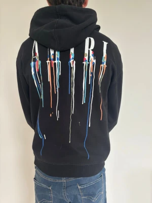 Svart hoodie med färgstänk från Amiri - Säljer en svart hoodie från Amiri med stor huva och känguruficka. På ryggen finns ett stort AMIRI-tryck med färgstänk i blått, orange, vitt och grönt som rinner ner. Framtill finns ett mindre tryck med samma design. Perfekt för dig som gillar streetwear och unik stil.
