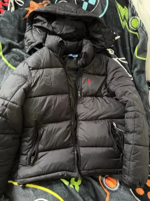 Svart pufferjacka med huva Polo Ralph Lauren - Svart pufferjacka från Polo Ralph Lauren med avtagbar huva och dragkedja framtill. Jackan har två stora fickor med dragkedja och den klassiska röda loggan broderad på bröstet. Tillverkad i glansigt, vadderat material som håller värmen jackan är storlek S men passar som XS