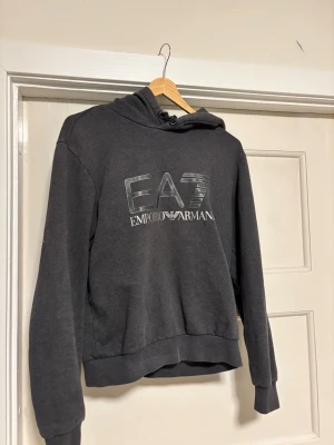 Mörkgrå hoodie EA7 Emporio Armani - Snygg mörkgrå hoodie från EA7 Emporio Armani med stor logga framtill. Klassisk modell med huva och dragsko, ribbade muddar vid ärmslut och nederkant. Perfekt för en avslappnad och trendig stil.