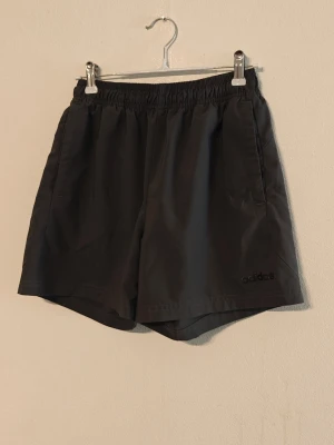 Adidas träningsshorts - Snygga gråa shorts från Adidas med elastisk midja och diskret logga på benet. Klassisk passform och tillverkade i lätt syntetmaterial som passar perfekt till träning eller chill. Två sidofickor för det viktigaste. Storlek L för barn.