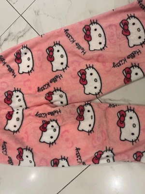 Rosa Hello Kitty mjukisbyxor - Supergosiga rosa mjukisbyxor med Hello Kitty-motiv och elastisk midja. Byxorna är i fleece och har ett mönster med vita Hello Kitty-huvuden och röda rosetter över hela tyget. Perfekta för mysiga hemmakvällar!