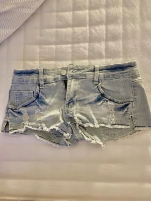 Ljusblå slitna jeansshorts - Säljer ett par ljusblå jeansshorts med slitna detaljer och fransig kant. Shortsen har fem fickor, bälteshällor och knappstängning framtill. Perfekta för varma dagar och ger en avslappnad vibe.
