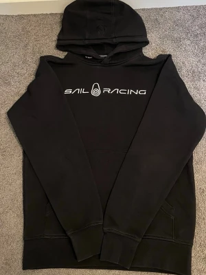 Svart hoodie från Sail Racing - Säljer en svart hoodie från Sail Racing med vit logga tryckt över bröstet. Hoodien har huva och en klassisk känguruficka framtill. Perfekt för dig som gillar stilren och sportig design. Materialet känns mjukt och bekvämt.