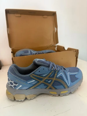 Blå sneakers från Asics - Snygga blå sneakers från Asics med meshpaneler och detaljer i grått och gult. Skorna har en sportig design med robust sula och klassiska Asics-loggan på sidan. Perfekta för dig som gillar streetstyle och bekväma skor.