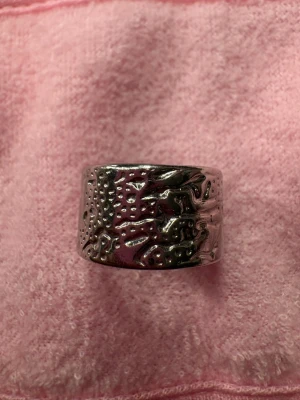 Bred silverfärgad ring med struktur - Cool bred ring i silverfärgad metall med unik, hamrad och ojämn yta som ger en rå och trendig känsla. Perfekt för dig som gillar statement-smycken och vill sticka ut med en annorlunda accessoar. Ringen har en rund form och är helt utan stenar eller pärlor.