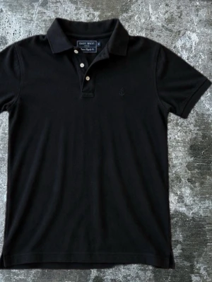 Svart piké från East West - Klassisk svart pikétröja från East West med krage och två knappar framtill. Diskret broderad ankare på bröstet ger en marin känsla. Perfekt till jeans eller shorts för en clean look.