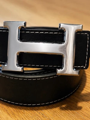 Svart skärp med silverfärgat H-spänne - Stilrent svart bälte i skinn med kontrasterande vita sömmar och ett stort, silverfärgat H-format spänne. Perfekt accessoar för att lyfta din outfit med en klassisk och modern touch.