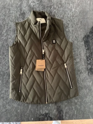 Olivgrön quiltad dunväst Burberry - Snygg olivgrön dunväst från Burberry med quiltat mönster. Väldigt skön och bra kvalité. Säljer pga av att den inte passade mig.