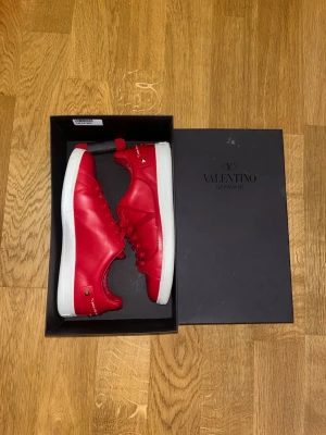 Valentino sneakers - Snygga röda sneakers från Valentino Garavani i slät läderlook med vita sulor. Skorna har matchande röda skosnören, diskret V-logga på sidan och små gulddetaljer vid hälen. Perfekta för dig som vill sticka ut med stilrena och exklusiva sneakers.