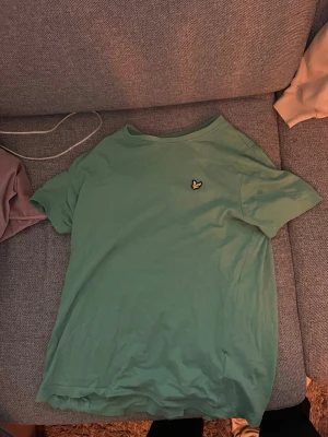 Lyle & Scott t shirt - Snygg grön t-shirt med korta ärmar och rund hals. På bröstet sitter en liten broderad logga i form av en gul fågel. Perfekt till jeans eller shorts för en avslappnad stil.