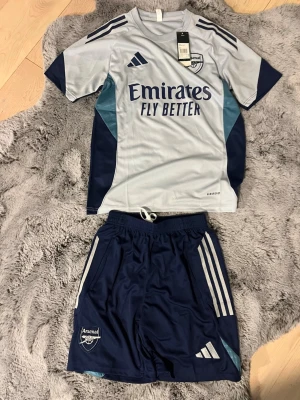 Arsenal träningsset i blått och grått - Snyggt Arsenal träningsset från Adidas med ljusgrå t-shirt och mörkblå shorts. Tröjan har klubbmärke, sponsortryck och klassiska Adidas-ränder på axlarna. Shortsen är mörkblå med klubbmärke och vita ränder. Perfekt för fotbollsträning eller gym. Storlek S, passar M bra