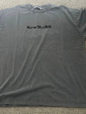 Grå Acne Studios t-shirt - Säljer en grå t-shirt från Acne Studios med svart logga på bröstet. T-shirten har en klassisk passform med rund hals och korta ärmar. Perfekt för en clean och stilren look.