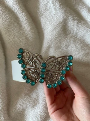 Vitt bälte med fjärilsspänne  - Snyggt vitt bälte med ett stort fjärilsspänne i silver dekorerat med turkosa stenar. Bältet har en bred rem och spännet har detaljerade mönster som gör det till en riktig statement-accessoar.