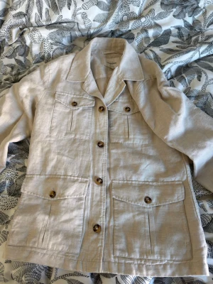 Beige linnéovershirt   - Stilren beige overshirt i linnematerial med fyra stora fickor framtill och knappar i trälook. Klassisk krage och långa ärmar med knappdetaljer vid ärmslut. Perfekt för en avslappnad men snygg look. Nypris 1599 mitt pris 649