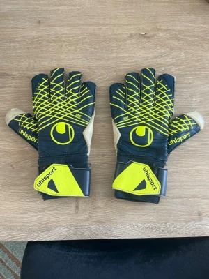 Uhlsport målvaktshandskar  - Säljer ett par svarta och gula målvaktshandskar från Uhlsport med justerbara kardborreband vid handleden. Handskarna har ett coolt mönster på ovansidan och slitstarka handflator i beige latex för bra grepp. Perfekta för fotbollsträning eller match!