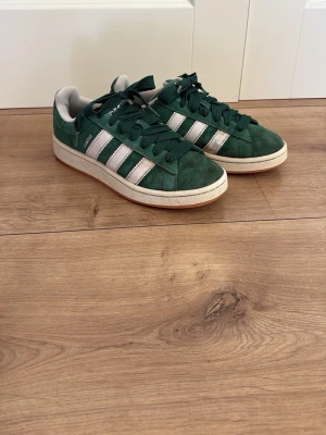 Gröna Adidas Campus sneakers - Snygga gröna Adidas Campus sneakers i mocka med vita detaljer och klassiska tre ränder på sidorna. Skorna har vit sula och gröna skosnören. Adidas-logga syns på hälen och sidan. Perfekta för en streetwear-look.