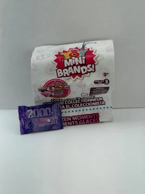 Mini Brands samlarleksak och guide - Mini Brands samlarleksak med tillhörande samlarguide. Förpackningen är vit med färgglad logga och text, och innehåller en liten lila leksaksförpackning märkt 2000G. Perfekt för dig som gillar miniatyrer och samlarprylar.