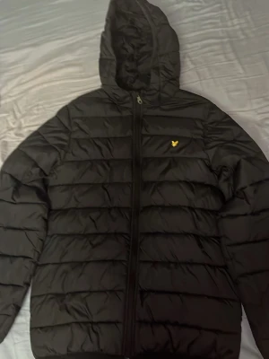 Svart lyle and scott pufferjacka med huva - Säljer en svart lyle and Scott pufferjacka med huva och dragkedja framtill. Jackan har ett quiltat mönster och en liten gul logga på bröstet. Perfekt för kalla dagar och riktigt skön att bära. Materialet är syntet och jackan har en modern, avslappnad passform. Pris and diskuteras vid snabb affär.