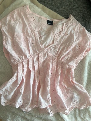 Blus från gina - Söt ljusrosa blus med broderade blommor och volangkant. Blusen har v-ringning, korta ärmar och smock i midjan för en feminin siluett. Perfekt för vår och sommar när du vill ha något luftigt och romantiskt.