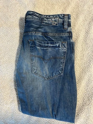 Blå raka jeans med slitningar - Säljer ett par klassiska blå jeans med raka ben och snygga slitningar. Jeansen har fem fickor, normal midja och kontrastsömmar. Perfekta för en avslappnad och trendig look. Materialet är denim i bomull.