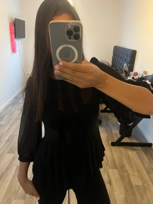Svart blus med volang och transparenta ärmar - Snygg svart blus med volang nertill och transparenta långärmade ärmar. Blusen har en figurnära passform och är gjord i ett lätt, tunt material som ger en elegant look. Perfekt att styla med jeans eller kjol för en trendig outfit.