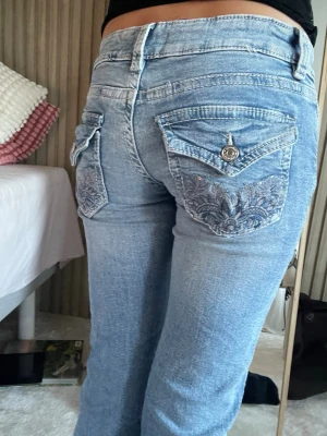 Ljusblå bootcut jeans med broderi - Säljer ett par ljusblå bootcut jeans med snygga broderade bakfickor och ficklock med knapp. Jeansen har låg midja och klassisk femficksdesign. 