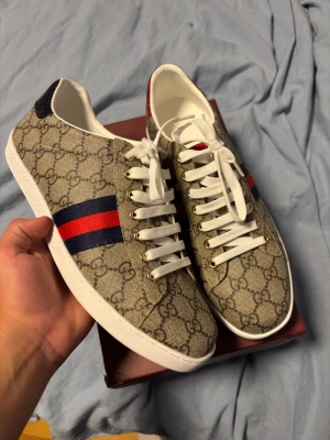 Gg skor  - Snygga gg sneakers i beige canvas med klassiskt GG-mönster och blå/röda ränder på sidan. Vita snören och sula, samt detaljer i mörkblått på hälen. Ikonisk logga under sulan. Perfekta för dig som vill sticka ut med stil.