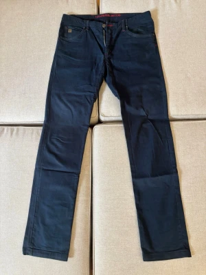 Mörkblå jeans från Hansen & Jacob - Snygga mörkblå jeans från Hansen & Jacob med raka ben och klassisk femficksmodell. Jeansen har detaljer i rött vid dragkedjan och knappstängning med logotyp. Materialet känns mjukt och stretchigt för extra komfort.