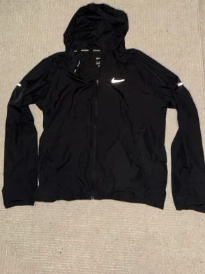 Svart Nike vindjacka med huva - Snygg svart vindjacka från Nike med huva och dragkedja framtill. Jackan har reflekterande detaljer på ärmarna och bröstet samt två sidofickor. Perfekt för blåsiga dagar och sportiga outfits.