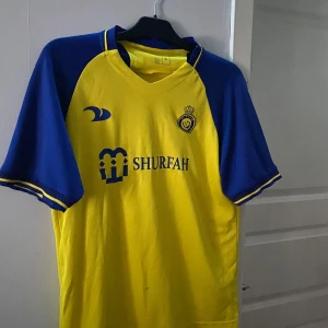 Al Nassr Ronaldo tröja - Säljer en gul och blå fotbollströja med korta ärmar från Shurfah. Tröjan har ett tryck med 'Ronaldo' och nummer 7 på baksidan. Perfekt för fotbollsfans som vill visa sitt stöd.