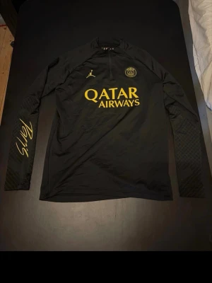 Svart PSG träningströja  - Snygg svart långärmad träningströja från PSG x Jordan med guldfärgade detaljer och loggor. Tröjan har halv dragkedja, Paris-tryck på ärmen och Qatar Airways som sponsor på bröstet. Perfekt för fotbollsträning eller streetwear. Passar S och M!