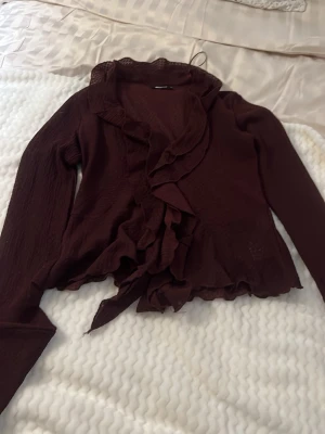Vinröd volangblus från Gina Tricot - Supersnygg vinröd blus från Gina Tricot med långa ärmar och volangdetaljer längs krage och framkant. Blusen har en lätt skrynklig struktur och knytband i midjan. Perfekt för dig som vill ha en trendig och feminin look.