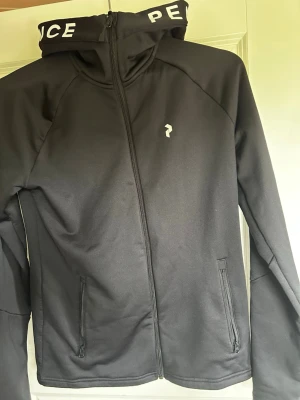 Peak performance hoodie/zip - Säljer en svart hoodie med dragkedja och huva. På huvan står det peak performance i vitt tryck och framtill finns en liten vit peak performance logga. Hoodien har två fickor med dragkedja och är gjord i ett mjukt, stretchigt material. Perfekt för en avslappnad och stilren look. Tröjan är i storlek M och är endast använd 1 gång. Pris kan diskuteras.