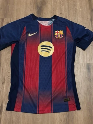Barcelona matchtröja Nike blå/röd - Säljer en officiell FC Barcelona matchtröja från Nike i blått och rött med klubbmärke, Spotify-logga och UNHCR-tryck. Tröjan har korta ärmar, rund hals och är tillverkad i ett lätt, ventilerande material perfekt för fotboll eller träning.