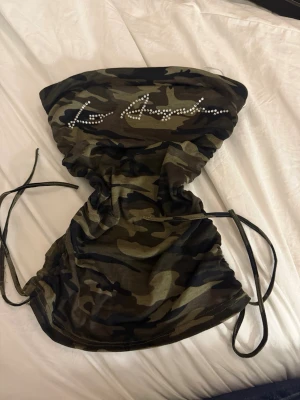 Camouflage bandeau tubtopp med snörning - Säljer en bandeau tubtopp i camouflage med snörning i sidorna och glittriga stenar framtill. Toppen är axelbandslös och har en tight passform, perfekt för en edgy och trendig stil.
