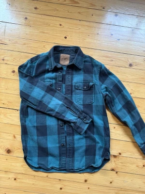 Rutig blå och svart skjorta - Snygg rutig skjorta från American Eagle i blått och svart. Skjortan har klassisk krage, bröstficka med knapp och långa ärmar. Perfekt för en avslappnad och cool stil. Tillverkad i mjuk bomull för extra komfort.