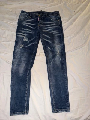 Blå dsquared2 jeans - Säljer ett par blå ds2 jeans med slitningar och distressed detaljer på både fram- och baksida. Jeansen har klassisk femficksmodell, låg midja och dragkedja med knapp. Perfekta för en avslappnad och trendig look.
