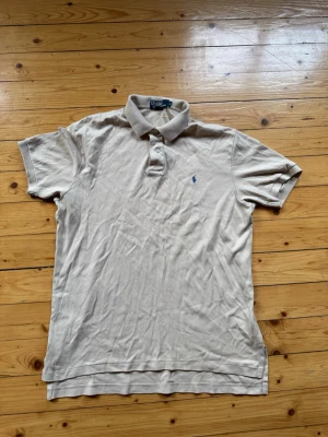 Beige pikétröja från Polo Ralph Lauren - Klassisk beige pikétröja från Polo Ralph Lauren med krage, knappar och broderad logga på bröstet. Tillverkad i mjuk bomull och har korta ärmar. Perfekt för en avslappnad och stilren look.