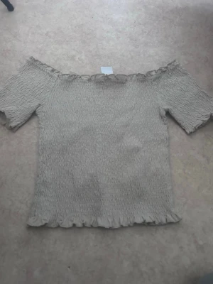Beige offshoulder topp med volang - Säljer en beige offshoulder topp med korta ärmar och volangdetaljer vid halsringning och nederkant. Toppen har en smockad, stretchig struktur som ger en tight passform och en söt, feminin look. Perfekt att styla med jeans eller kjol.
