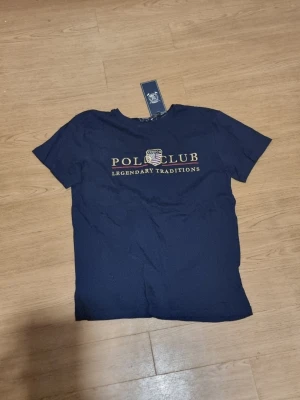 Mörkblå Polo Club t-shirt - INSTABOX!!! Snygg mörkblå t-shirt från United Polo Club med tryck i vitt och guld på bröstet. Klassisk passform och rund halsringning, perfekt för en stilren och avslappnad look.