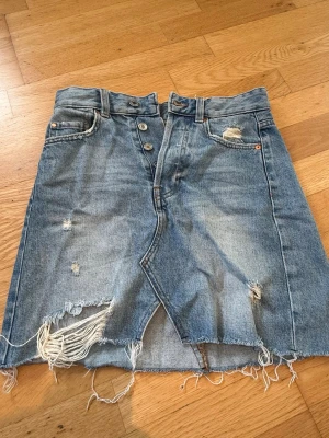 Blå jeanskjol med slitningar - Säljer en blå jeanskjol med slitna detaljer. Kjolen har knappar framtill, och fransig nederkant.