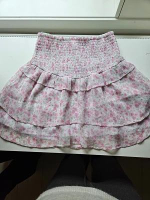  volangkjol - Supersöt kort kjol med blommigt mönster i rosa och vitt. Kjolen har bred smock i midjan och dubbla volanger för en extra flowy känsla. Perfekt för vår och sommar när du vill ha en lätt och luftig look. Skriv för mer information och bilder!