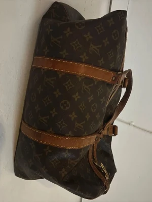 Louis Vuitton monogram travelbag - Vintage travelbag från Louis Vuitton i klassiskt brunt monogrammönster med detaljer i ljusbrunt läder och guldfärgade dragkedjor. Väskan har dubbla handtag och ikoniska LV-loggor på både tyg och metalldetaljer. Perfekt för dig som gillar lyxiga accessoarer. 